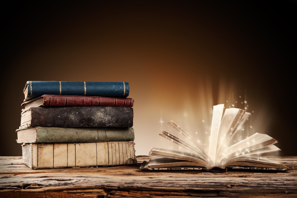 A stack of vintage books next to an open magical book emitting light and sparkles on a wooden surface.