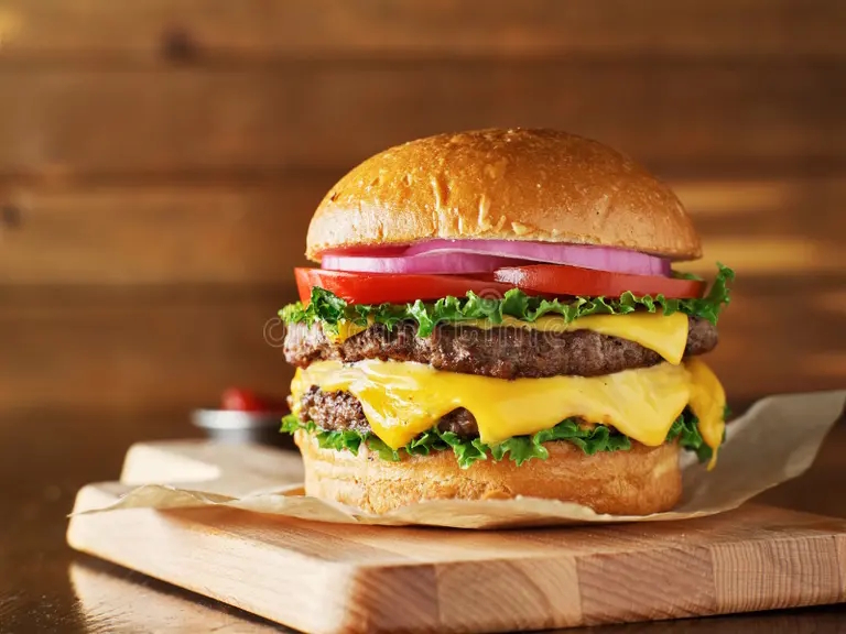 Close-up of a gourmet hamburger with two beef patties, melted cheddar cheese, lettuce, tomato, red onion, and a toasted bun, served on a wooden cutting board.