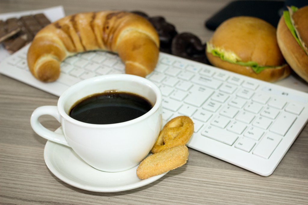 A cup of black coffee on a white saucer next to a small cookie, with a croissant and two sandwiches in the background, placed on a keyboard.
