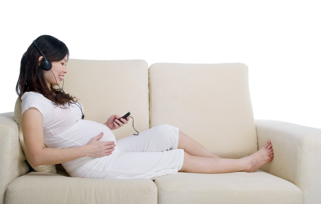 Pregnant woman sitting on a couch, listening to music through headphones while looking at a device.