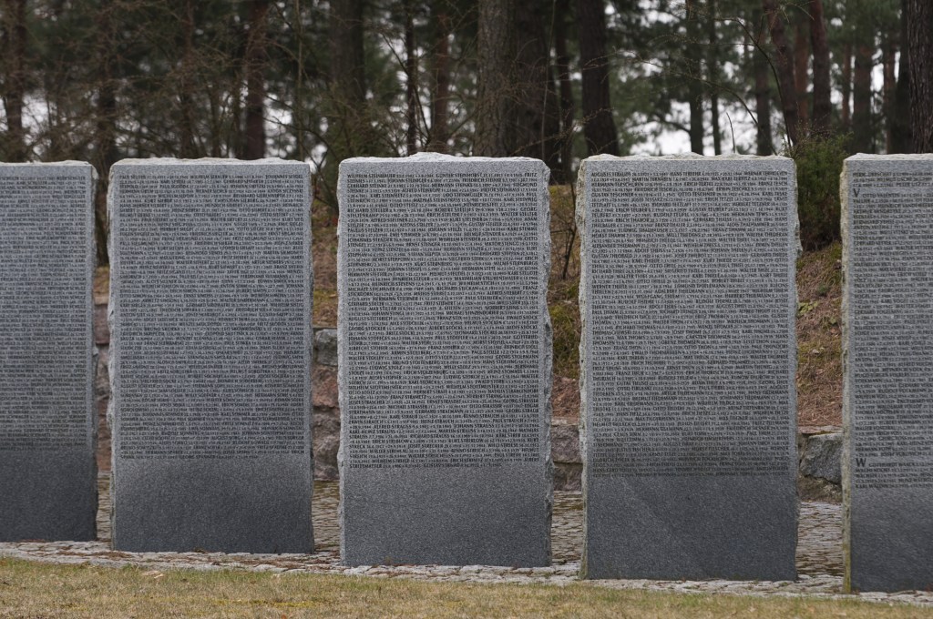 A series of tall granite memorial stones inscribed with text, set in a natural outdoor environment surrounded by trees.